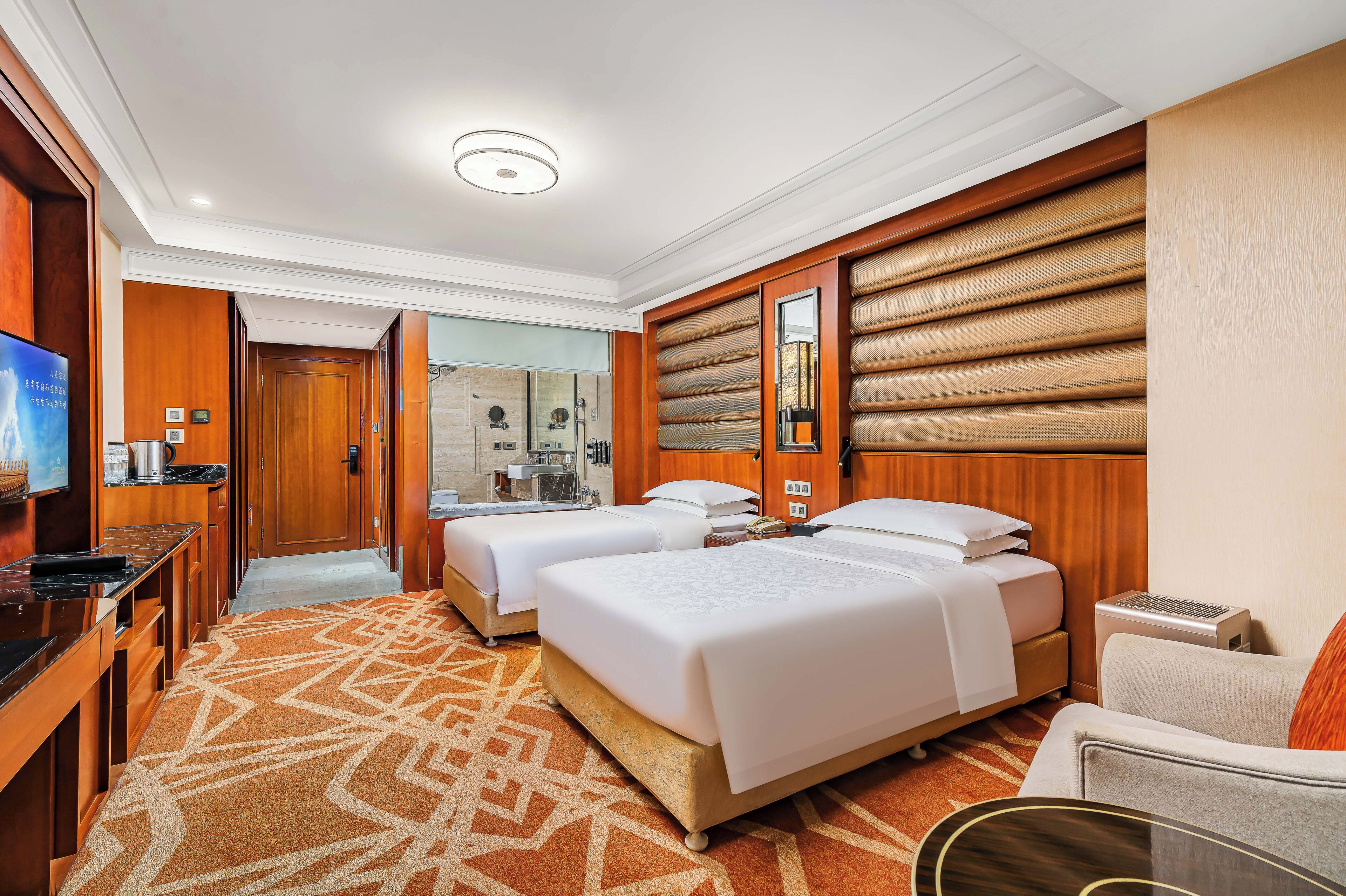 A hotel room with two beds, a TV, a desk, and a bathroom visible through an open door.