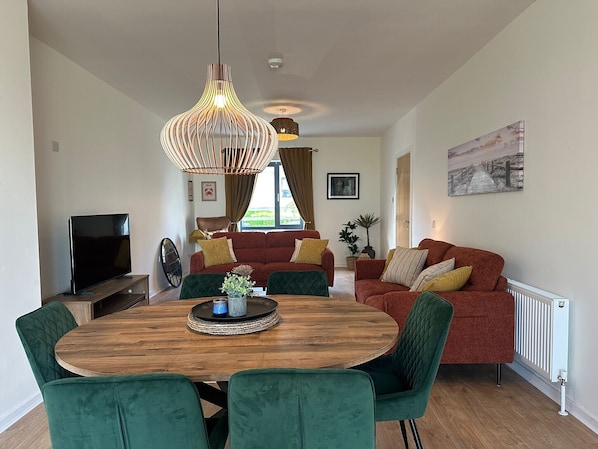 Ein modernes Wohnzimmer mit einem Holzesstisch, grünen Polsterstühlen, einem roten Sofa mit gelben Kissen und einer hängenden Leuchte.