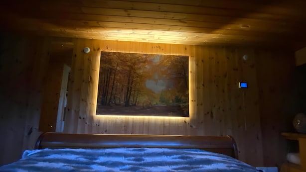 Et værelse med en seng, et billede af en skov på væggen og en lille hylde med en lampe.