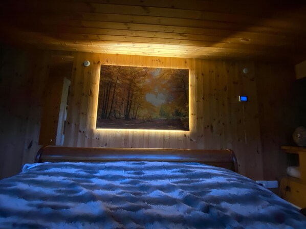 Una habitación con una cama, un cuadro colgado en la pared de un bosque, y una pequeña estantería con una lámpara.