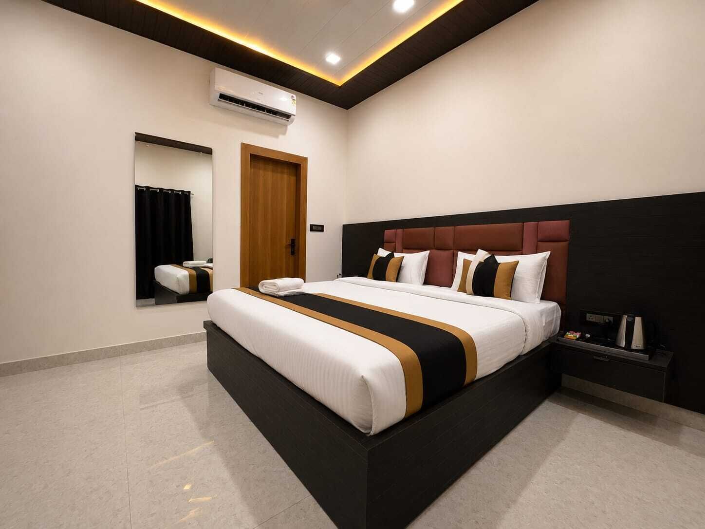 A modern hotel bedroom with a large bed, a bedside table, an air conditioning unit, and a mirror.