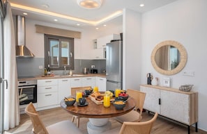 A modern kitchen with white cabinetry, a stainless steel refrigerator, and a dining area with a wooden table set for breakfast. - Grand Villa with Private Pool (Kefalonia)