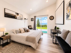 A bedroom with a bed, bedside tables, a desk, a bench, and a large window with a view of greenery.