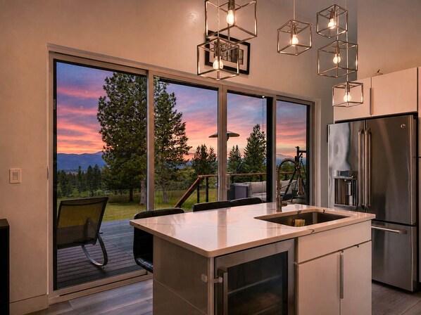 A modern kitchen with a large island, stainless steel appliances, and a view of a sunset through floor-to-ceiling windows.