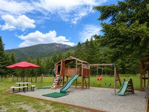 A playground with a wooden playhouse, slides, picnic tables, and a red umbrella.