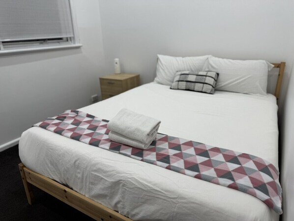 A single bed with a white mattress, a grey blanket, and a patterned throw. There is a wooden nightstand with a white lamp beside the bed. - Family Home for 12 in Werribee (Werribee)