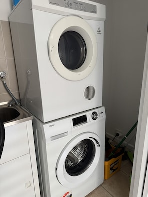 Lavadora y secadora apiladas, con panel de control, manguera de entrada de agua y una cesta de ropa amarilla.