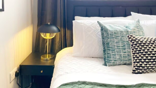 Ein ordentlich bezogenes Bett mit weißen Leinentüchern, einem dunklen Kopfteil und gemusterten Kissen. Eine Nachttischlampe mit schwarzem Lampenschirm steht auf einem dunklen Nachttisch.