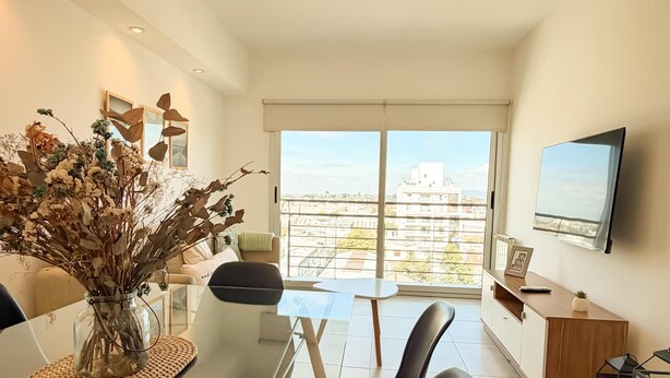 Ein modernes Wohnzimmer mit einem Glastisch, schwarzen Stühlen, einer Vase mit getrockneten Blumen, einem Sofa, einem Fernseher und Blick auf die Stadt durch große Fenster.