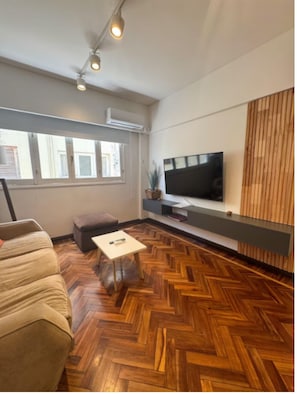 Ein modernes Wohnzimmer mit Fischgrätparkett, einer Couch, einem Couchtisch, einem Flachbildfernseher, der an einer Holzvertäfelung befestigt ist, und einem Fenster mit Jalousien.