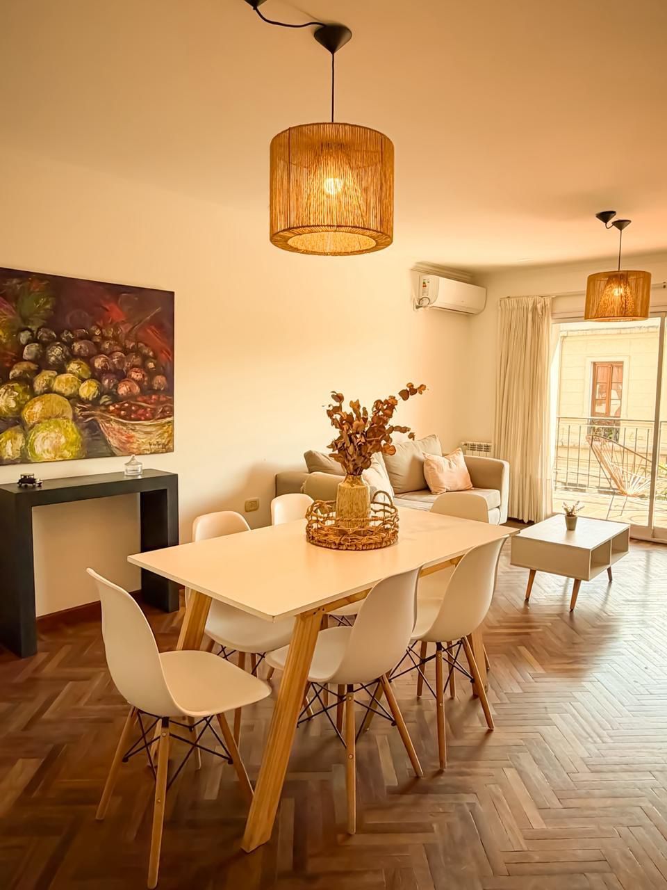 A dining area with a wooden table and chairs, a hanging light fixture, and a painting on the wall.