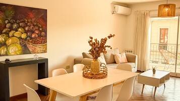 A dining area with a wooden table and chairs, a hanging light fixture, and a painting on the wall.