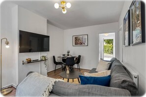 A modern living room with a sofa, a dining table, and a TV mounted on the wall. - Cosy 1 Bedroom Bungalow w Garden in Stepney Green (London)