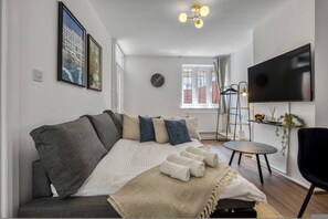 A modern living room with a sofa, a TV, a clock, and a window. - Cosy 1 Bedroom Bungalow w Garden in Stepney Green (London)