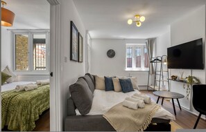 A modern living room with a sofa, a TV, and a small table. - Cosy 1 Bedroom Bungalow w Garden in Stepney Green (London)