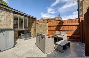 A patio with wicker furniture, a grey storage unit, and a brick wall. - Cosy 1 Bedroom Bungalow w Garden in Stepney Green (London)