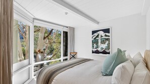 Ein Schlafzimmer mit großem Fenster, einem Bett mit Kissen, einem Sessel und einem gerahmten Bild an der Wand.
