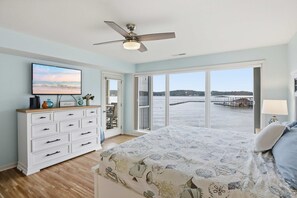 Ein Schlafzimmer mit Bett, Schminktisch, Deckenventilator, Fernseher und einem großen Fenster mit Blick auf einen Steg und Wasser.