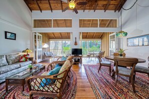 A spacious living area with a high ceiling, wooden beams, a television, wicker furniture, and a colorful rug.