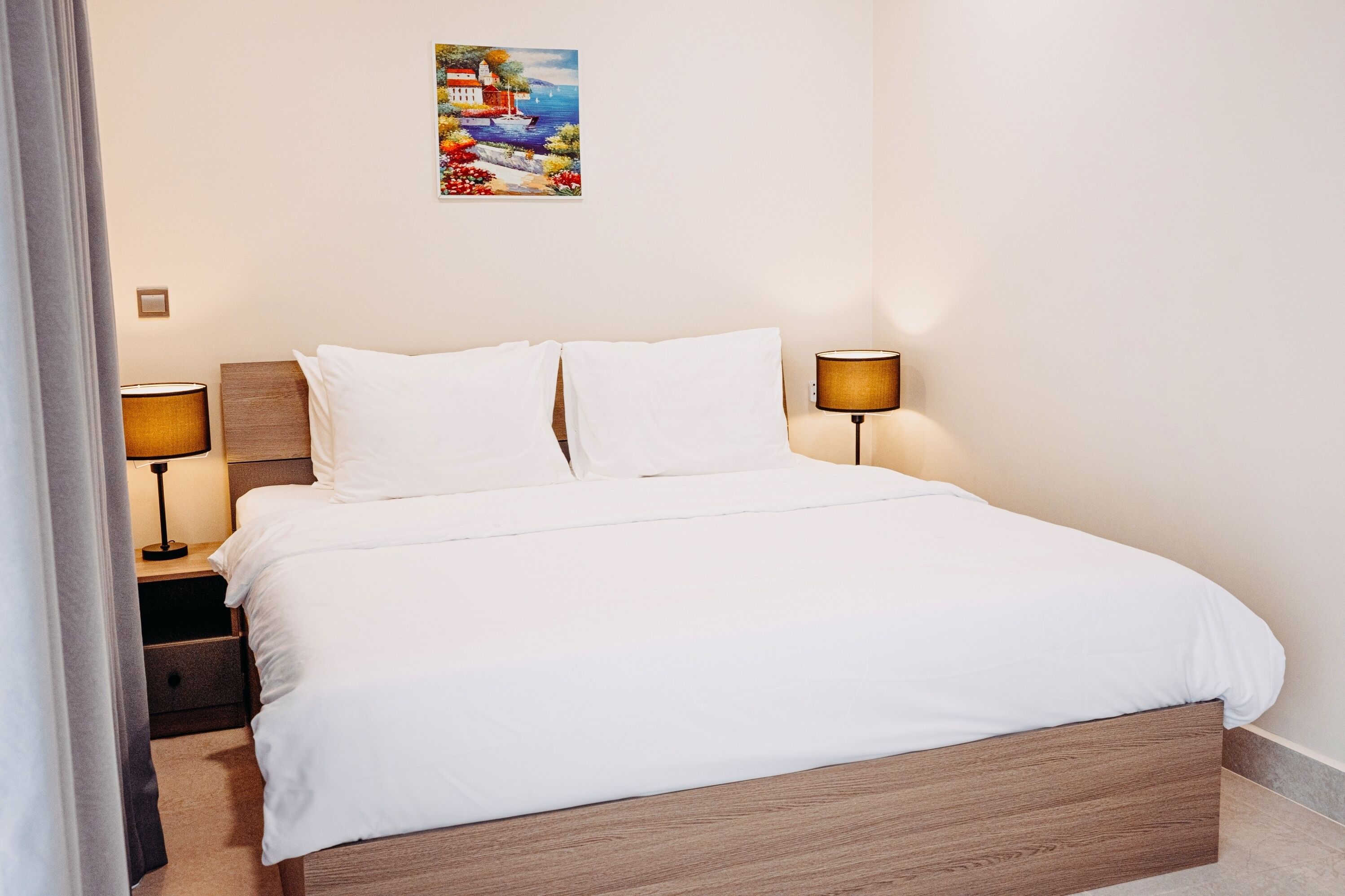 Una cama bien tendida con sábanas blancas, dos lámparas de noche, una mesita de noche y un cuadro colgado en la pared.