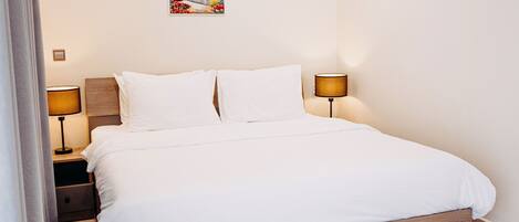 Una cama bien tendida con sábanas blancas, dos lámparas de noche, una mesita de noche y un cuadro colgado en la pared.