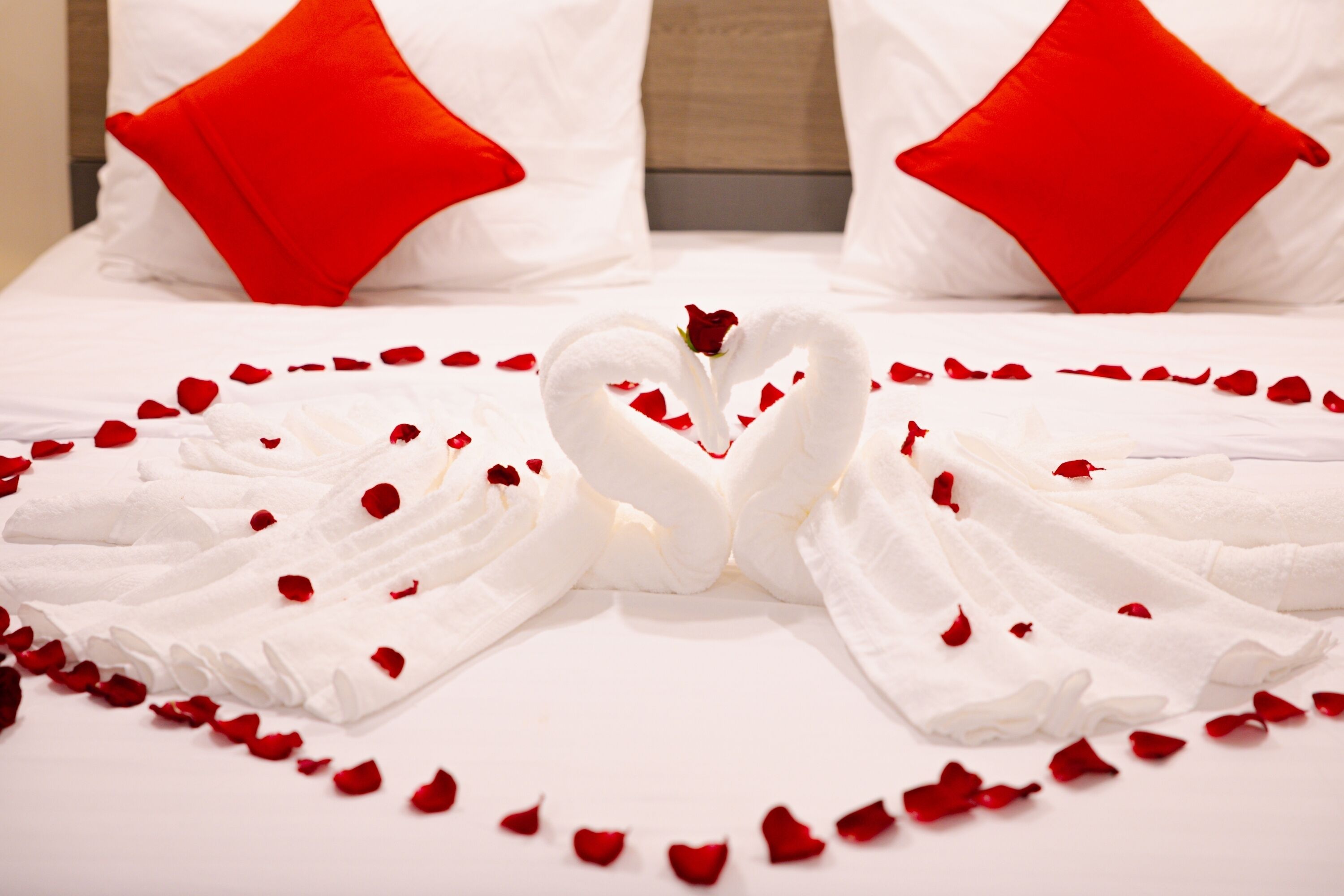 A bed with white linens, red pillows, and a heart-shaped arrangement of towels and rose petals.