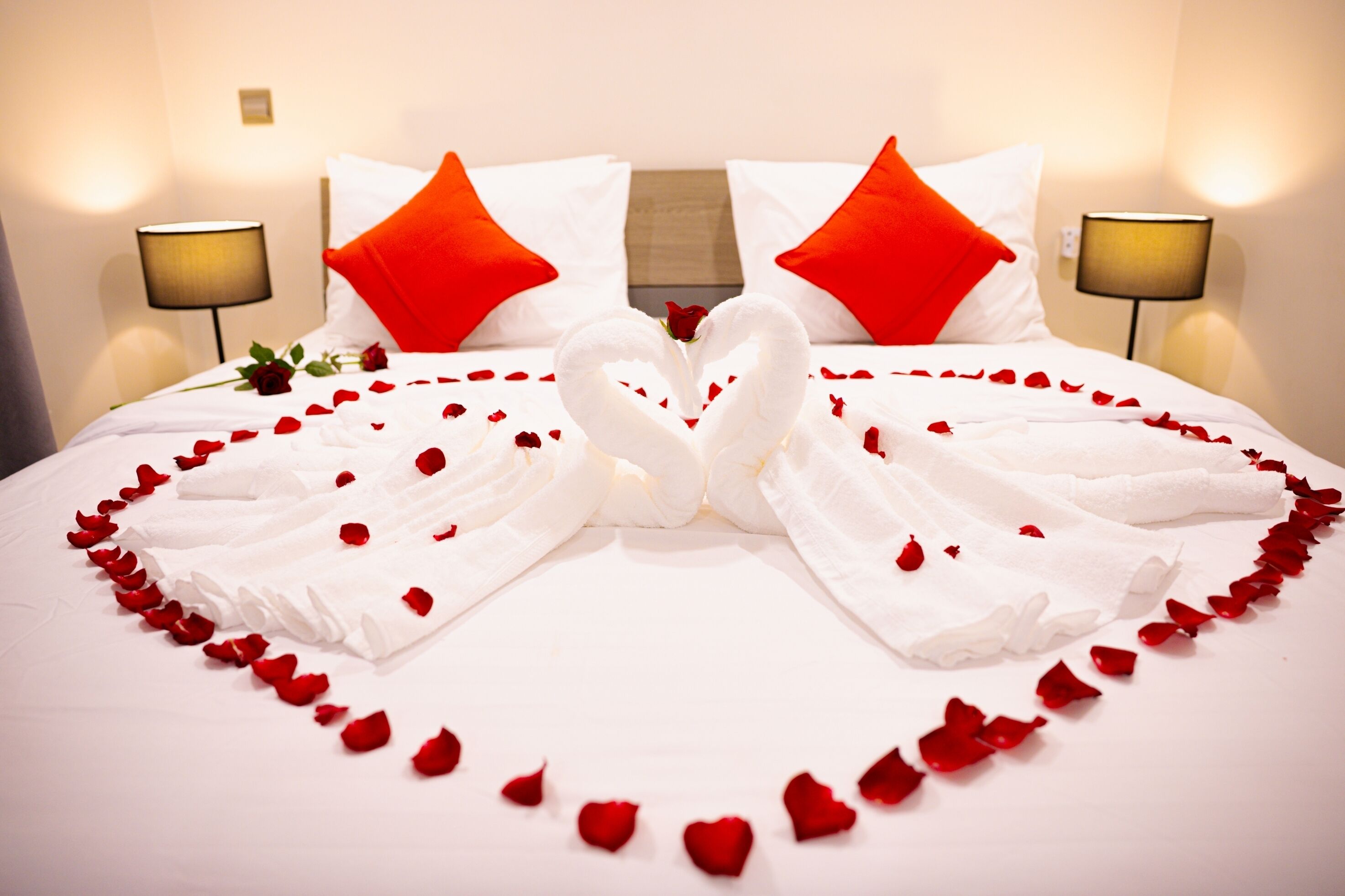 A bed with a heart-shaped towel arrangement and rose petals, white bedding, and red pillows.