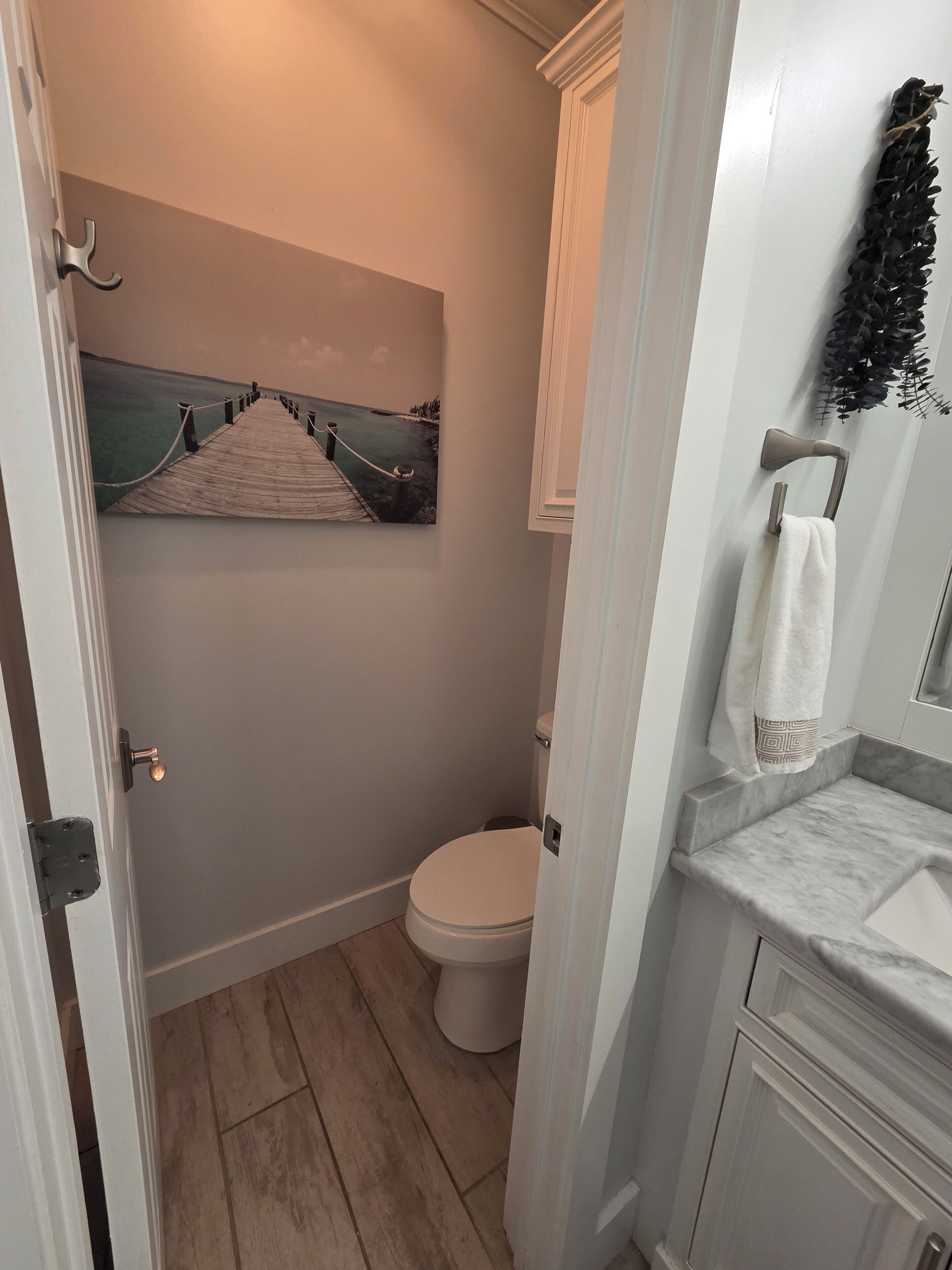 A bathroom with a wooden floor, a toilet, a sink with a marble countertop, and a painting of a pier.