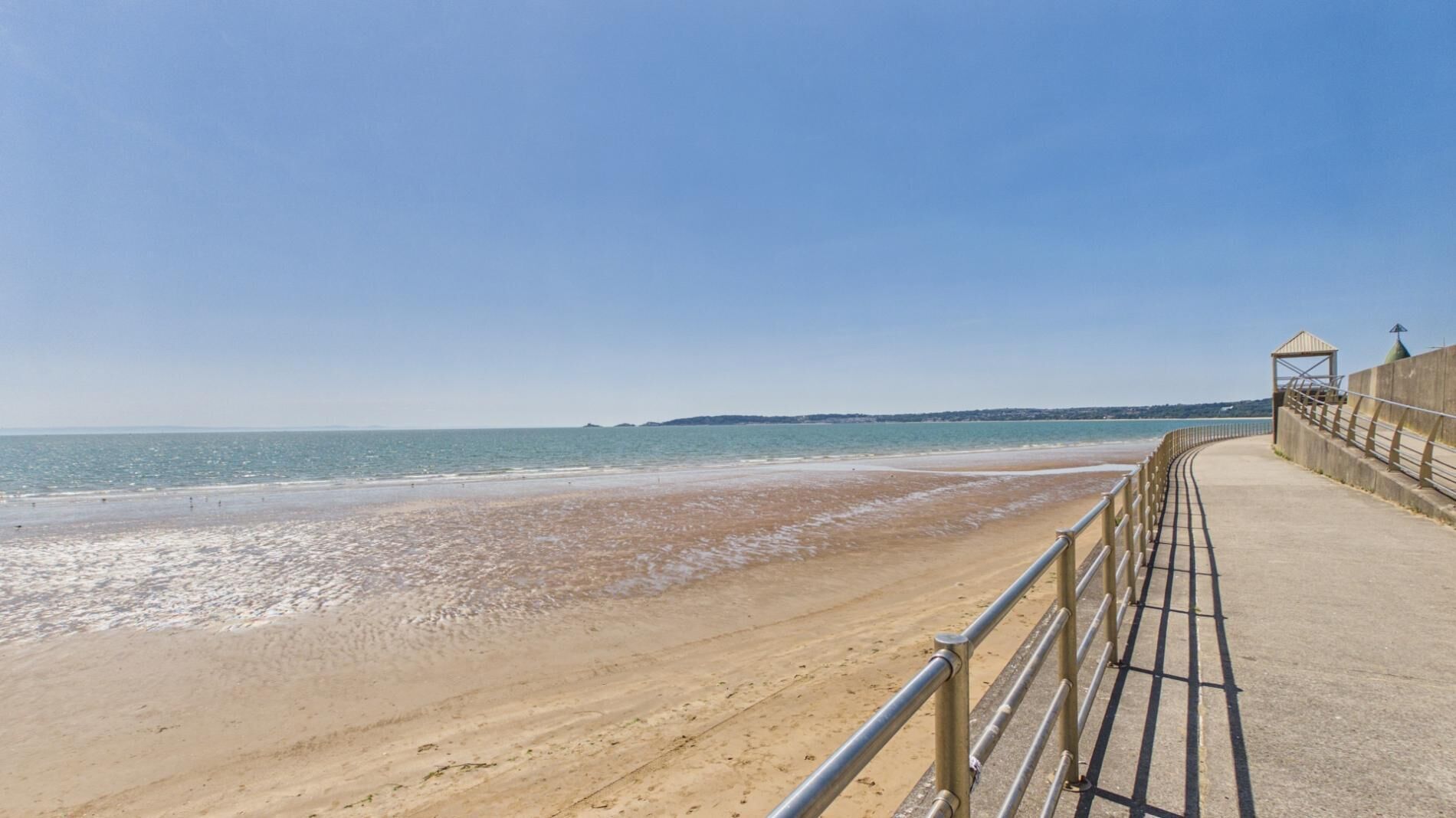 A coastal walkway with a metal railing, leading to a beach with gentle waves.