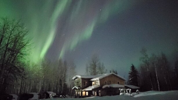 A house with a snow-covered roof, surrounded by trees, under a night sky with visible aurora borealis.