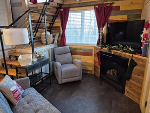 Una sala acogedora con chimenea, un sillón cómodo, un sofá, una mesita con lámpara y una escalera.