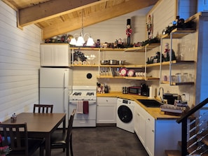 A compact kitchen with a stove, oven, microwave, and washing machine. There is a dining table with chairs, and shelves above the counter holding various kitchen items.