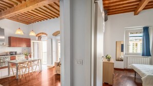 Una cocina moderna con vigas de madera, un comedor con mesa de vidrio y un dormitorio con ventana y radiador.