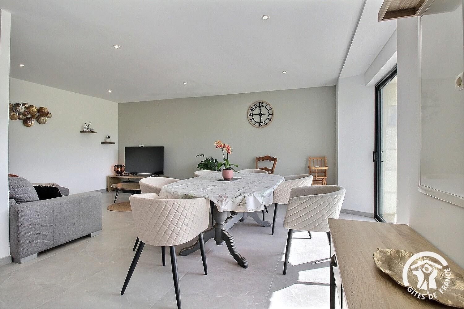 A modern living room with a dining area, a grey sofa, a TV, and a clock on the wall.