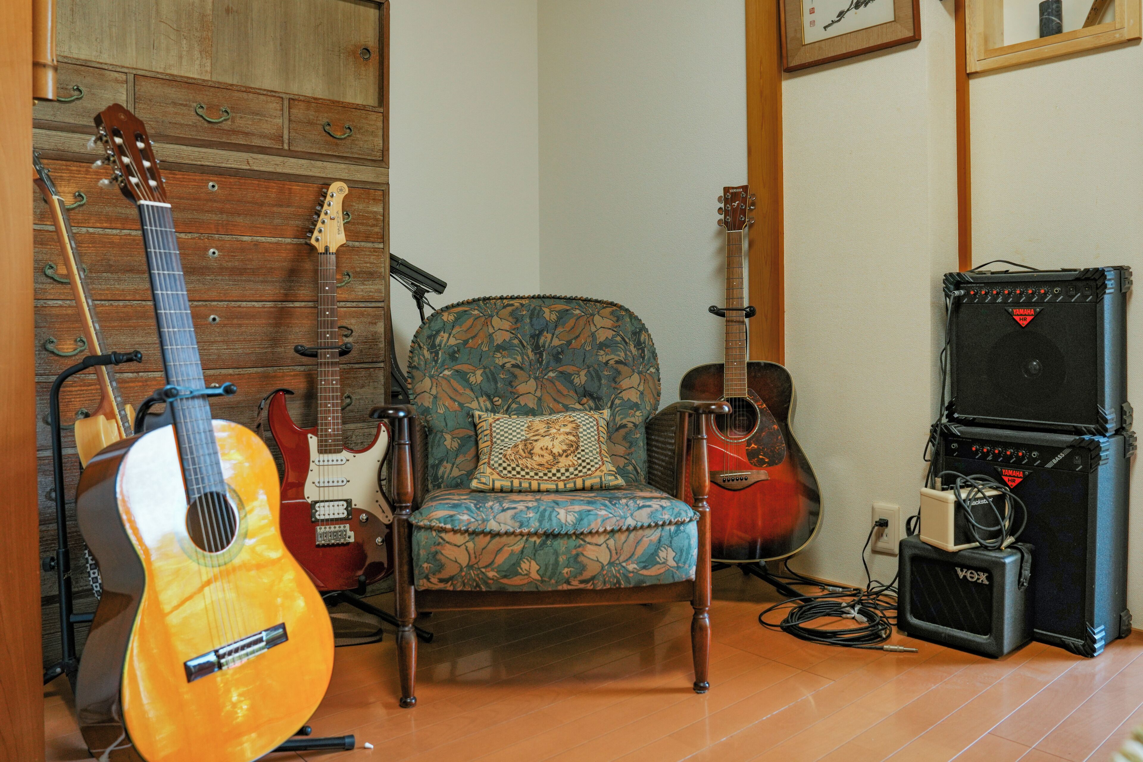 A room with a wooden wall, a patterned armchair, and various musical instruments including guitars and amplifiers.