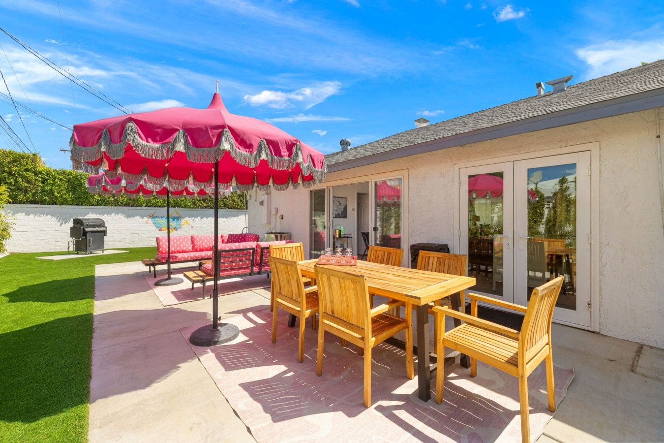 A patio with a wooden table and chairs, a red umbrella, and a barbecue grill.