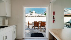 Une cuisine avec des meubles blancs, une table à manger en bois avec des chaises, et une vue sur un patio avec un bâtiment blanc et des palmiers.
