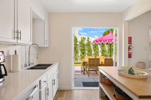 A modern kitchen with white cabinets, a stainless steel sink, and a dining area with wooden furniture. - Desert Divas Escape - Palm Springs (Palm Springs)