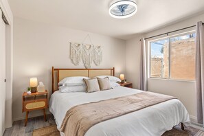 A bedroom with a bed, bedside tables, a wall-mounted macrame piece, a ceiling fan, and a window with curtains.
