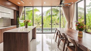 A modern kitchen with a large island, wooden ceiling, and large windows overlooking a garden. - Hometown Kem 4 BRS Pool Villa Hoi An (Hoi An)