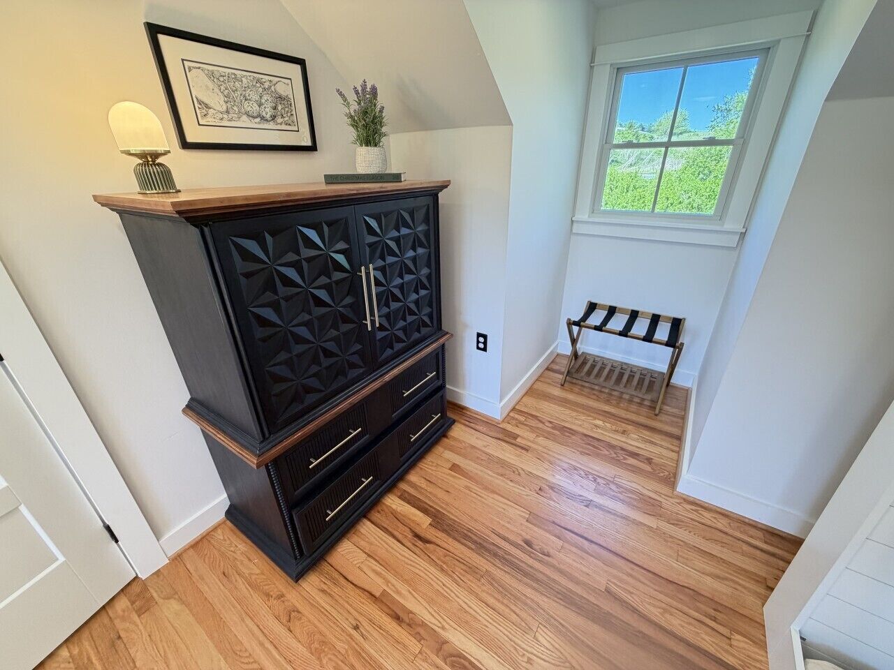 A room with a dark wooden cabinet, a framed picture, a small plant, and a window with a view of greenery.