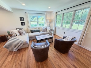 A living room with a sofa, ottoman, and coffee table, large windows with a view of greenery, and a lamp.