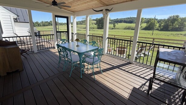 Eine überdachte Terrasse mit Tisch und Stühlen, einem Deckenventilator und Blick auf ein grünes Feld.
