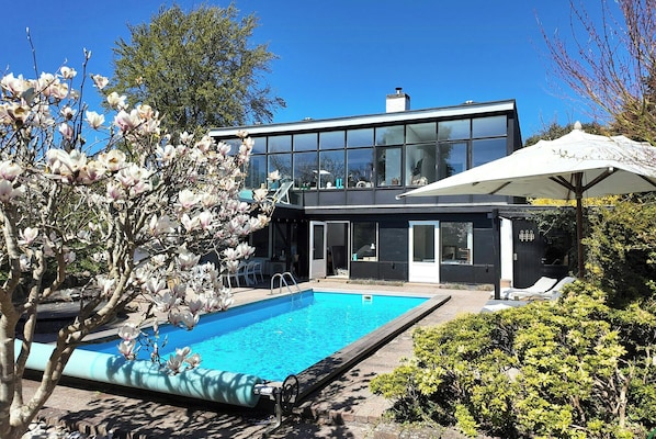 A modern house with a swimming pool, a covered patio, and a garden with flowering trees.