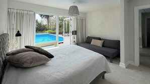 A bedroom with a large bed, a sofa, a view of a swimming pool, and a door leading to another room.