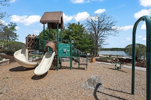 Ein Spielplatz mit Rutschen und Klettergerüsten, umgeben von Kiesflächen und Bäumen.
