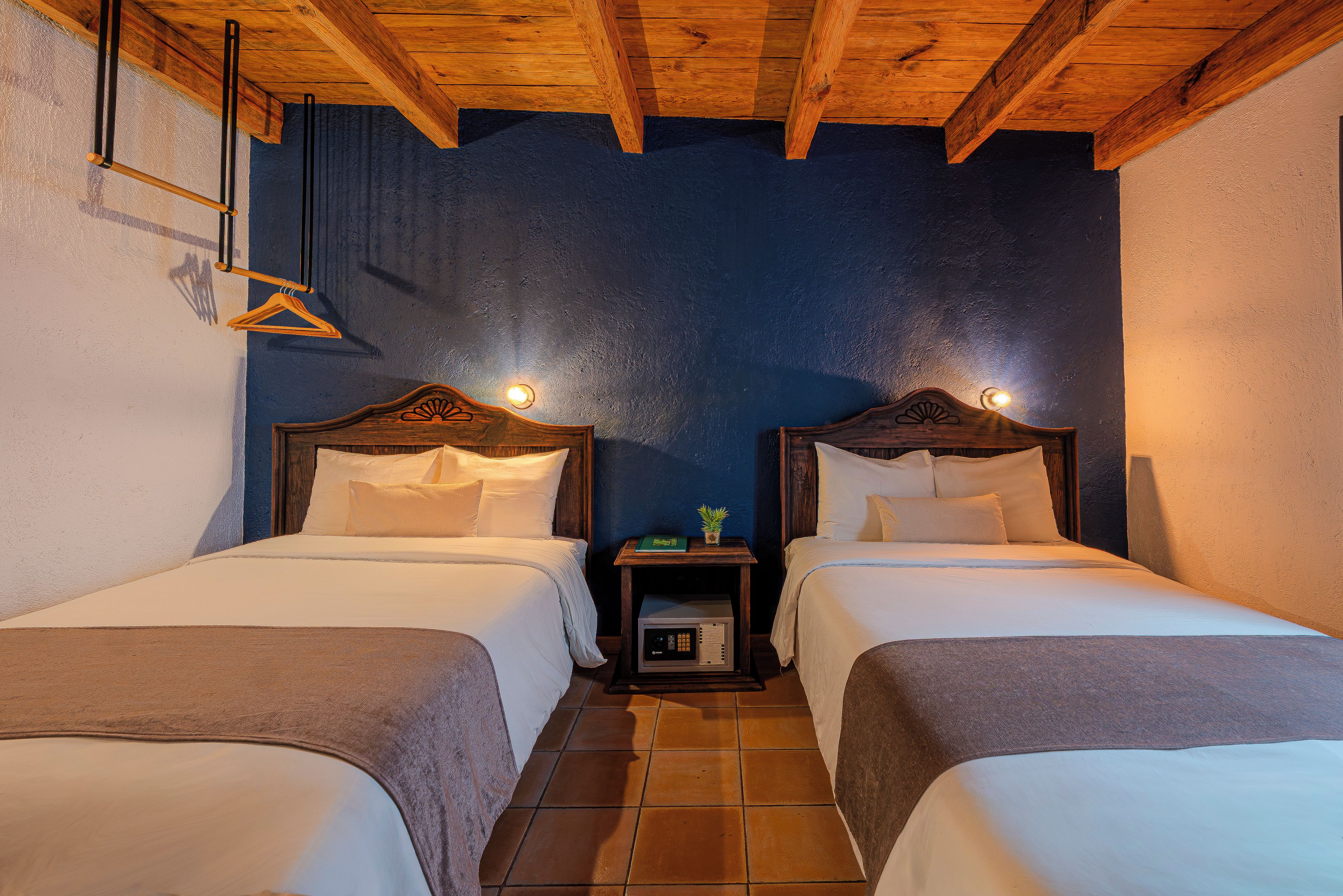 A room with two beds, wooden headboards, a small bedside table with a lamp, and a tiled floor.
