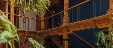 A wooden staircase leading to a balcony with hanging plants, a seating area with a sofa and cushions, and a tiled floor.