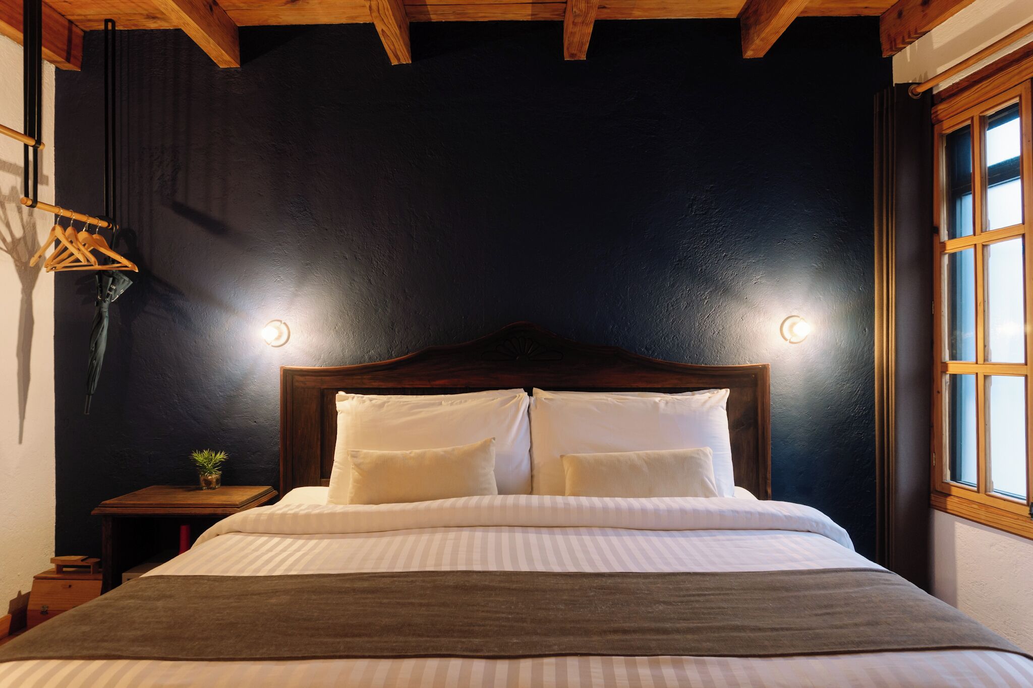 A bed with a wooden headboard, two bedside lamps, a small table with a plant, and a window with wooden frames.