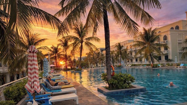 Un área de piscina con sillas de descanso, palmeras y un edificio al fondo.